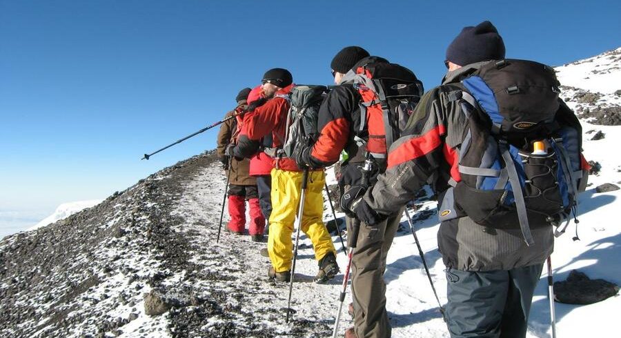 A group of hikers in colorful winter gear with backpacks and trekking poles traverse a snowy ridge on Kilimanjaro's Northern Circuit under a clear blue sky.