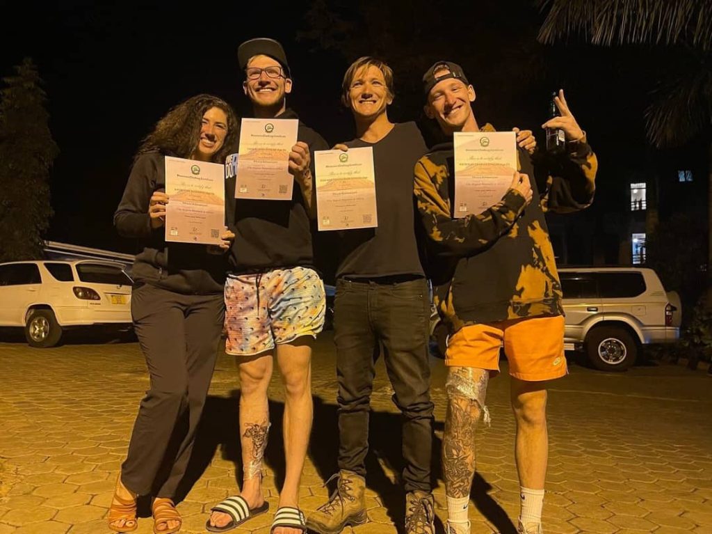 Group of four friends at night posing outdoors, each holding a certificate, wearing casual outfits including orange shorts and dark tops, illuminated by warm lighting, representing Climb Kilimanjaro and Tanzania Safaris events.