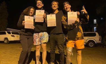 Group of four friends at night posing outdoors, each holding a certificate, wearing casual outfits including orange shorts and dark tops, illuminated by warm lighting, representing Climb Kilimanjaro and Tanzania Safaris events.