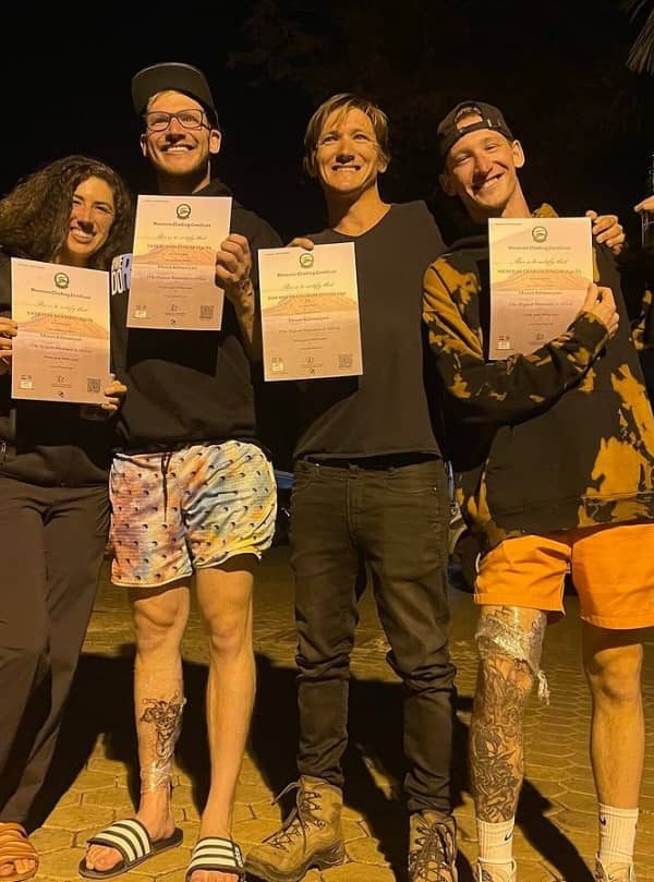 Group of four friends at night posing outdoors, each holding a certificate, wearing casual outfits including orange shorts and dark tops, illuminated by warm lighting, representing Climb Kilimanjaro and Tanzania Safaris events.