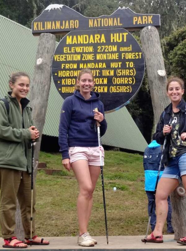 Three hikers pose near Kilimanjaro National Park sign in Tanzania, with trekking poles; green huts and forest backdrop, showcasing a misty outdoor trekking scene.