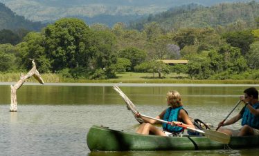 Two people wearing blue life jackets paddle a dark green canoe on a serene lake, surrounded by dense green trees and distant hills, capturing a tranquil outdoor scene.