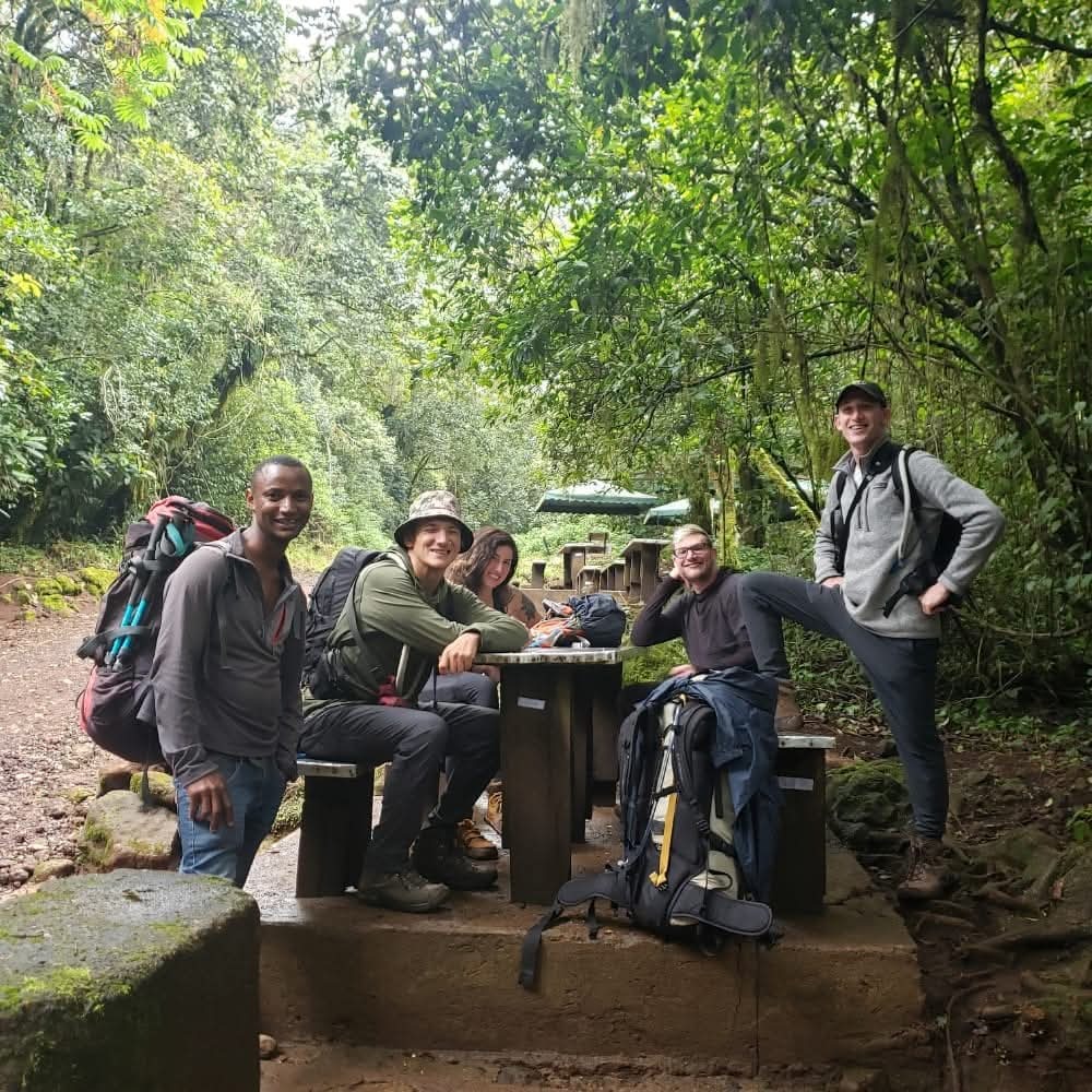 Marangu hikers gather at a wooden picnic table amid dense greenery, wearing backpacks and trekking gear as sunlight filters through the trees.
