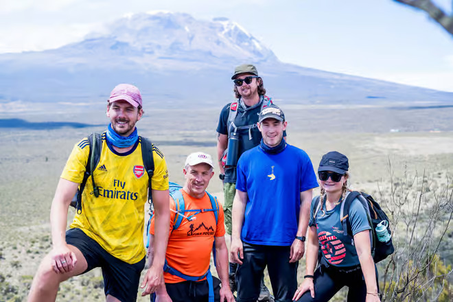 Group of five hikers posing on a desert trail with a distant mountain in the background.