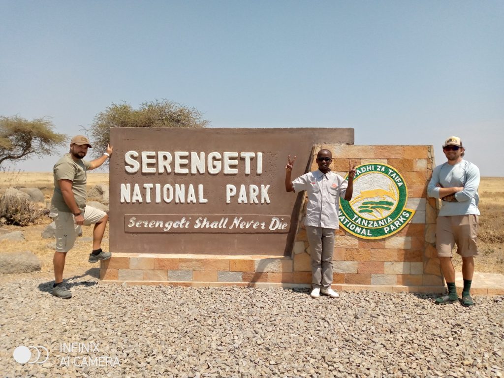 Three visitors pose by the Serengeti National Park sign in a dry, sunny landscape, two making peace signs.