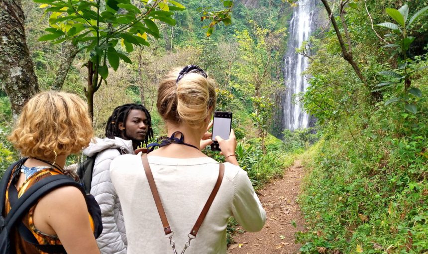 #image_title Three tourists on a dirt forest trail photograph a distant waterfall amid lush greenery.