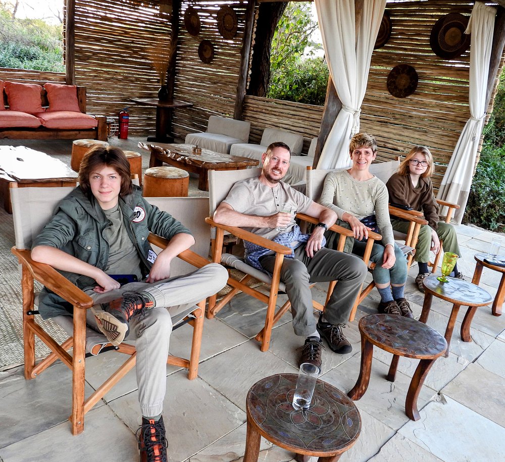 Five people sit in rustic outdoor lounge chairs under a shaded patio with wicker walls and white curtains on a sunny day.