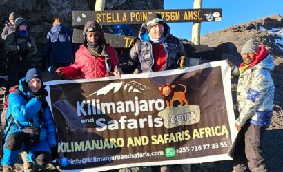 Group of hikers at the Kilimanjaro summit area holding a banner for Kilimanjar o and Safaris Africa, with a multi-line sign behind them against a clear blue sky and rocky terrain.