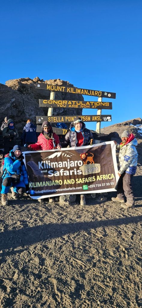 Group of hikers at the Kilimanjaro summit area holding a banner for Kilimanjar o and Safaris Africa, with a multi-line sign behind them against a clear blue sky and rocky terrain.
