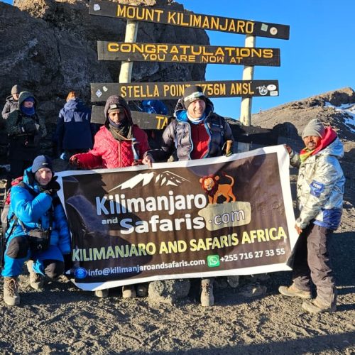 Group of hikers at the Kilimanjaro summit area holding a banner for Kilimanjar o and Safaris Africa, with a multi-line sign behind them against a clear blue sky and rocky terrain.