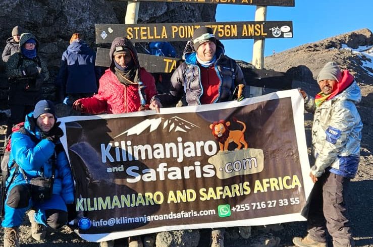 Group of hikers at the Kilimanjaro summit area holding a banner for Kilimanjar o and Safaris Africa, with a multi-line sign behind them against a clear blue sky and rocky terrain.
