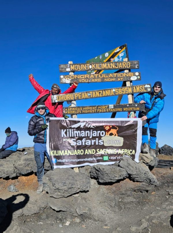 Group of climbers posing at Kilimanjaro summit sign, holding a banner for Kilimanjaro Safaris.