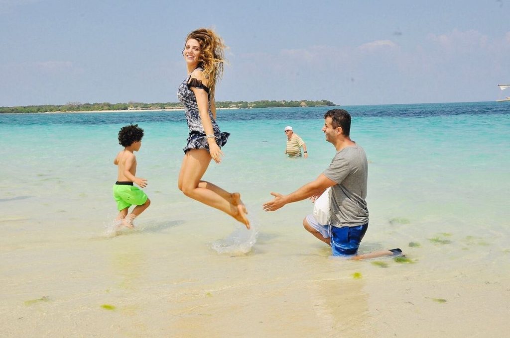 Woman in a patterned swimsuit mid-air, jumping in shallow turquoise water as a man in gray shirt and blue shorts reaches out nearby on a sunny beach.
