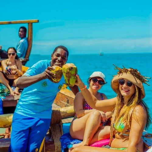 Friends on a tropical boat ride toasting with fresh coconuts against a bright blue sea and sky.
