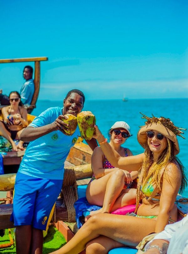 Friends on a tropical boat ride toasting with fresh coconuts against a bright blue sea and sky.