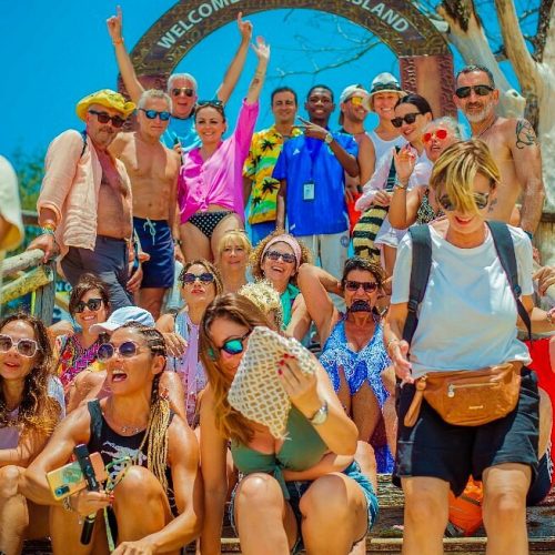 A large group of tourists posing on steps under a Welcome to Prison Island arch on a sunny day.