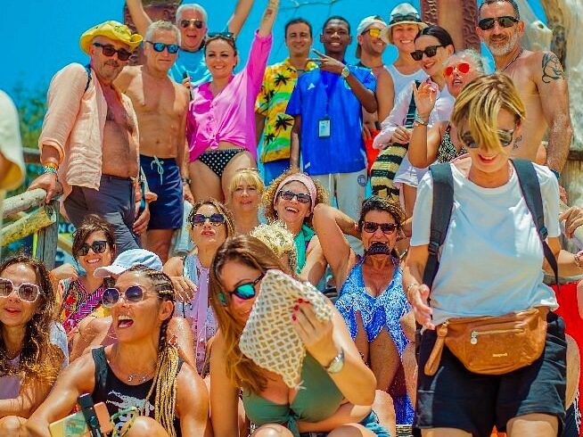 A large group of tourists posing on steps under a Welcome to Prison Island arch on a sunny day.