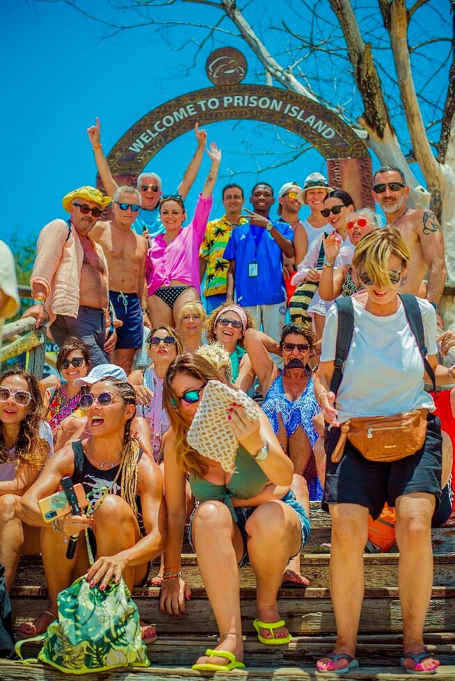 A large group of tourists posing on steps under a Welcome to Prison Island arch on a sunny day.