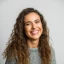 Portrait of a smiling woman with long curly hair looking at the camera, in a bright setting.