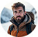 Portrait of a man with a beard wearing an orange outdoor jacket and backpack, with mountains in the background.