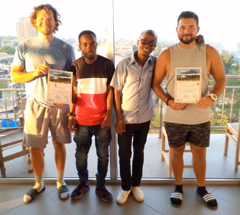 Four men pose on a bright balcony; two at the ends hold certificates and smile at the camera.