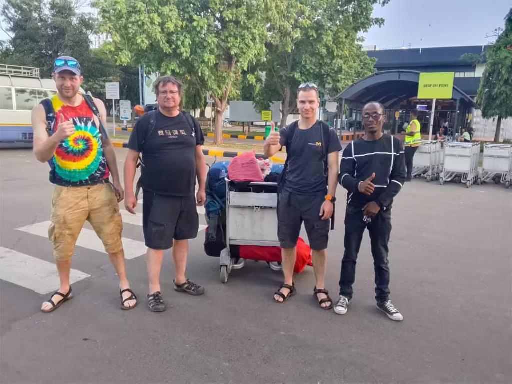 Group of four men posing with a luggage cart in a bus terminal area, giving thumbs up. One wears a tie-dye shirt and sandals; others wear casual shorts and sneakers. Trees and a station entrance are in the background.
