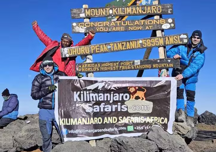 pavel and his frand depurture at Uhuru Peak with Climb Kilimanjaro and Safaris 2 | Climb Kilimanjaro and Tanzania Safari Three hikers pose at the Kilimanjaro summit sign, holding a Kilimanjaro Safaris banner under a clear blue sky. | Climb Kilimanjaro and Tanzania Safari
