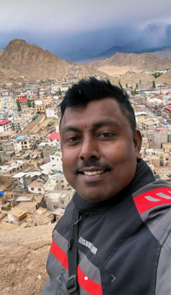 Sumeda Attanayaka 2 | Climb Kilimanjaro and Tanzania Safari | Climb Kilimanjaro and Tanzania Safari Self-portrait of a man smiling, with a hillside town and rocky mountains in the background. | Climb Kilimanjaro and Tanzania Safari | Climb Kilimanjaro and Tanzania Safari