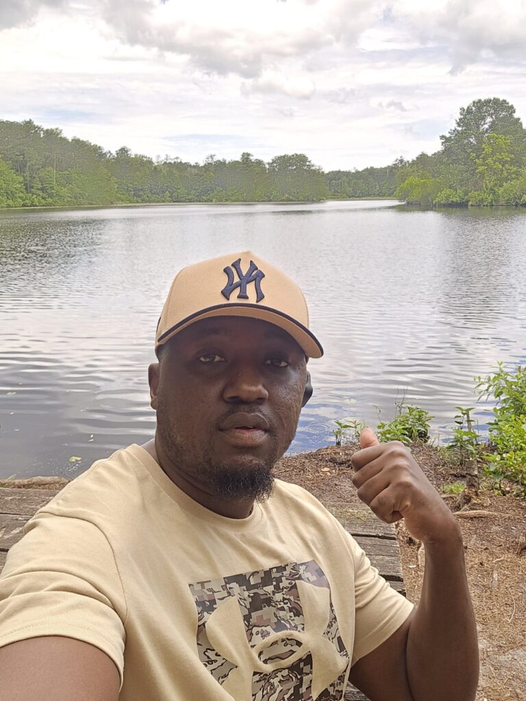 paul owiso | Climb Kilimanjaro and Tanzania Safari | Climb Kilimanjaro and Tanzania Safari Man wearing a beige NY Yankees cap takes a selfie by a lake with trees in the background, giving a thumbs-up gesture. | Climb Kilimanjaro and Tanzania Safari | Climb Kilimanjaro and Tanzania Safari