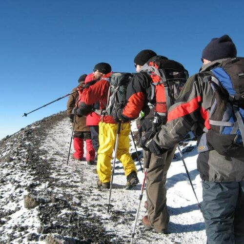 Group of hikers with backpacks and trekking poles walk along a snowy mountain ridge under a clear blue sky, wearing colorful jackets.