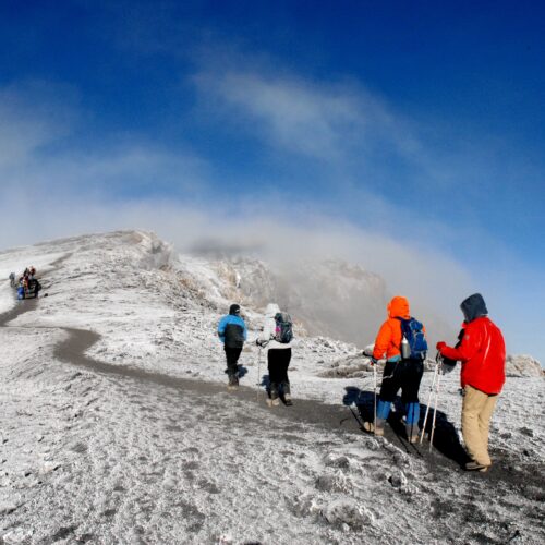 Group of hikers in bright jackets ascending a snow-covered mountain ridge under a clear blue sky.