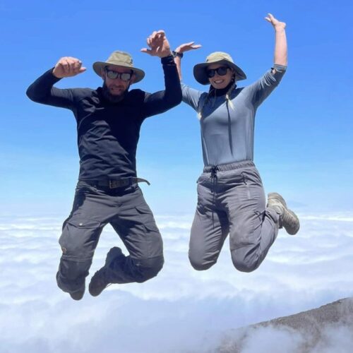 Two hikers in hats and sunglasses jump in the air above a sea of clouds on a mountain trail环境。