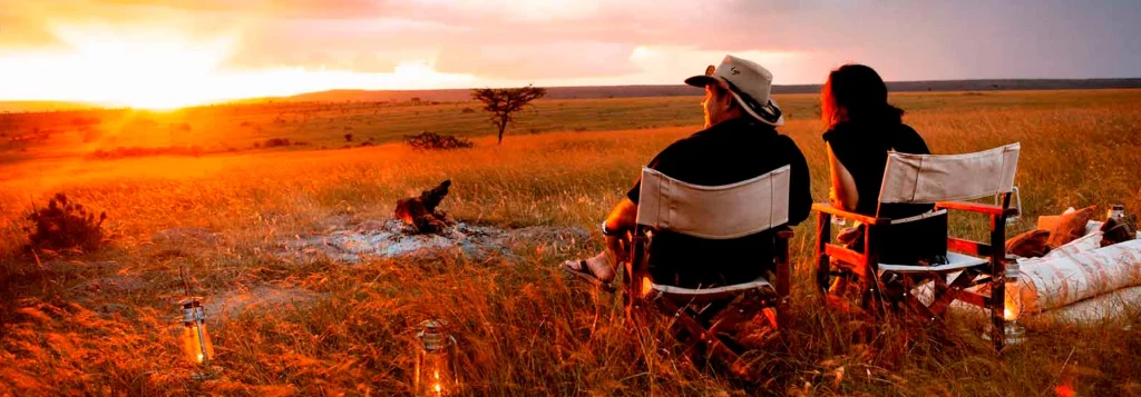 masai-mara | Climb Kilimanjaro and Tanzania Safari Two people sit in camping chairs around a small campfire at sunset on a wide grassy plain. | Climb Kilimanjaro and Tanzania Safari